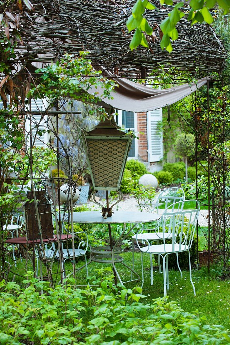 Zum Verkauf stehender Tisch und Gartenstühle aus weiss lackiertem Metall mit antiker Laterne unter Weidenpergola