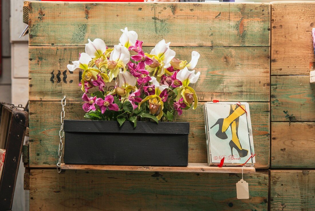 Schwarzer Schuhkarton mit verschiedenfarbigen Orchideen und Cleamtisblüten dekoriert neben gerahmtem Bild auf Holzablage, an Vintage Bretterwand montiert