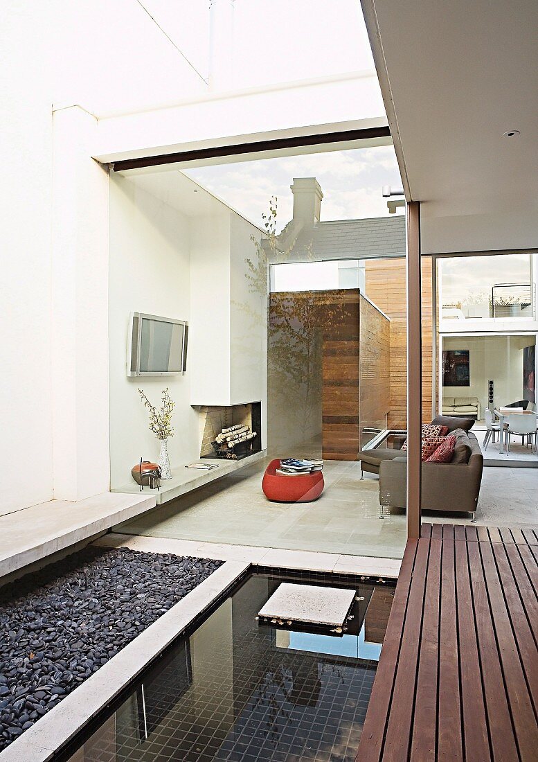 View from courtyard with gravel area, pool and wooden deck through frameless glass facade into seating area with fireplace and TV in modern interior