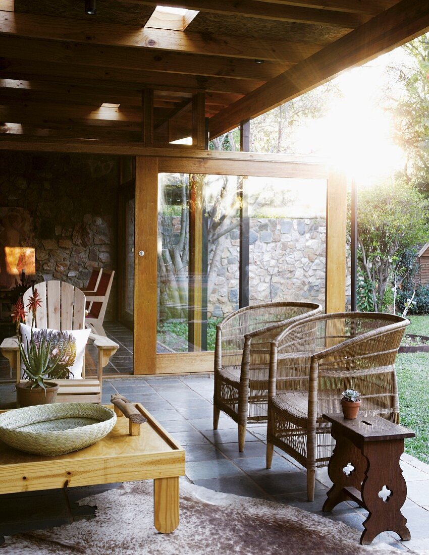 In das Haus integrierte, überdachte Terrasse mit Armlehnstühlen, Geflechtsesseln und Beistelltischen auf Tierfellteppich; Schiebetür vor dem Wohnraum im Hintergrund