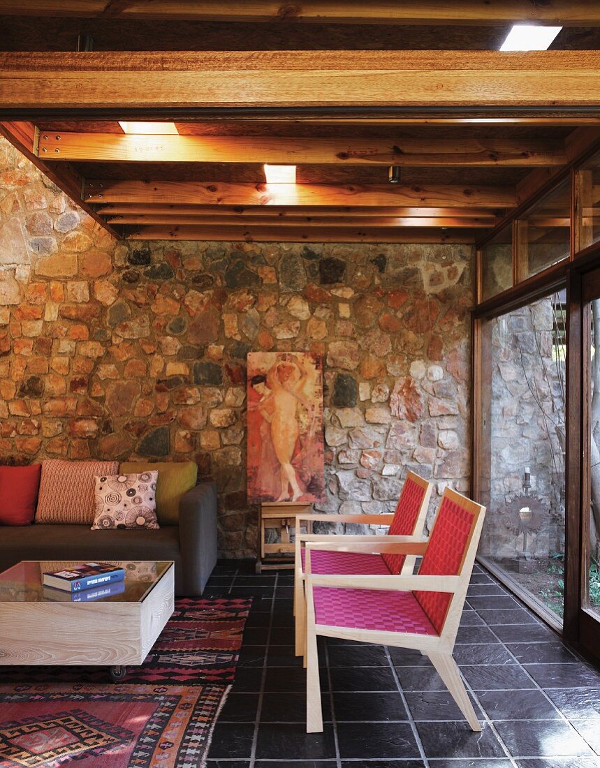 Sitzbereich mit pinkfarben bespannten Holzsesseln, Sofa und Kastentisch vor Natursteinwand; Glasschiebefront zum Garten und Holzdecke mit Lichtelementen