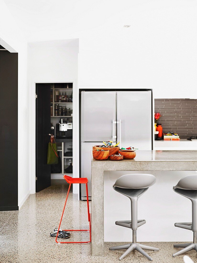Ausschnitt einer modernen Küche mit Kühlschrankelement und Hockern an Frühstücksbar aus Beton; seitlich Blick durch die offene Tür des Vorratsraumes
