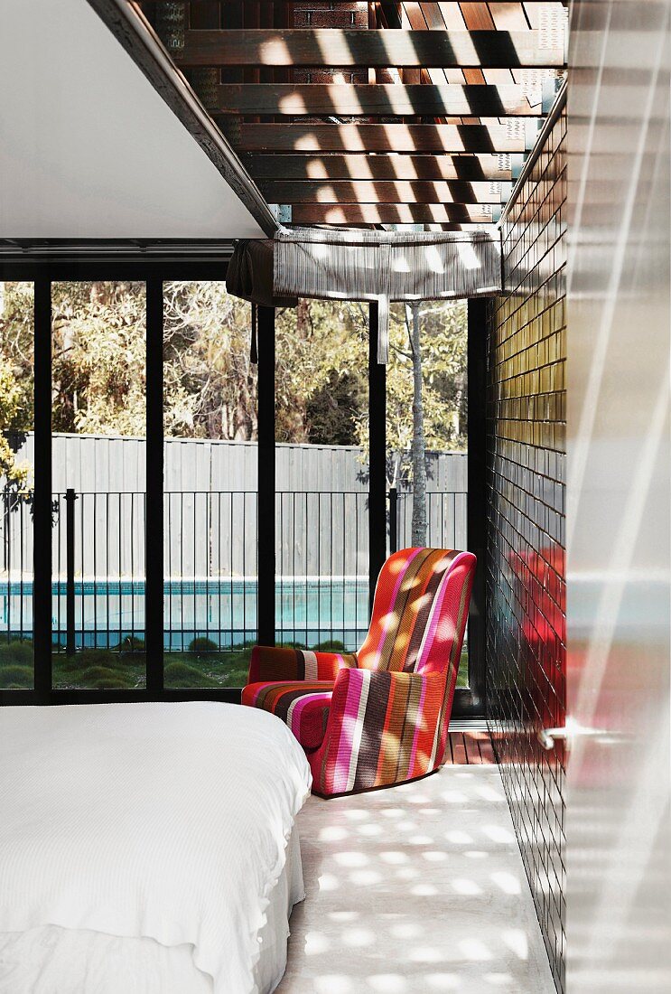 Double bed with white bed linen and wingback chair in colourful stripes in bedroom with pattern of light and shade falling through skylight; view of pool through glass wall in background