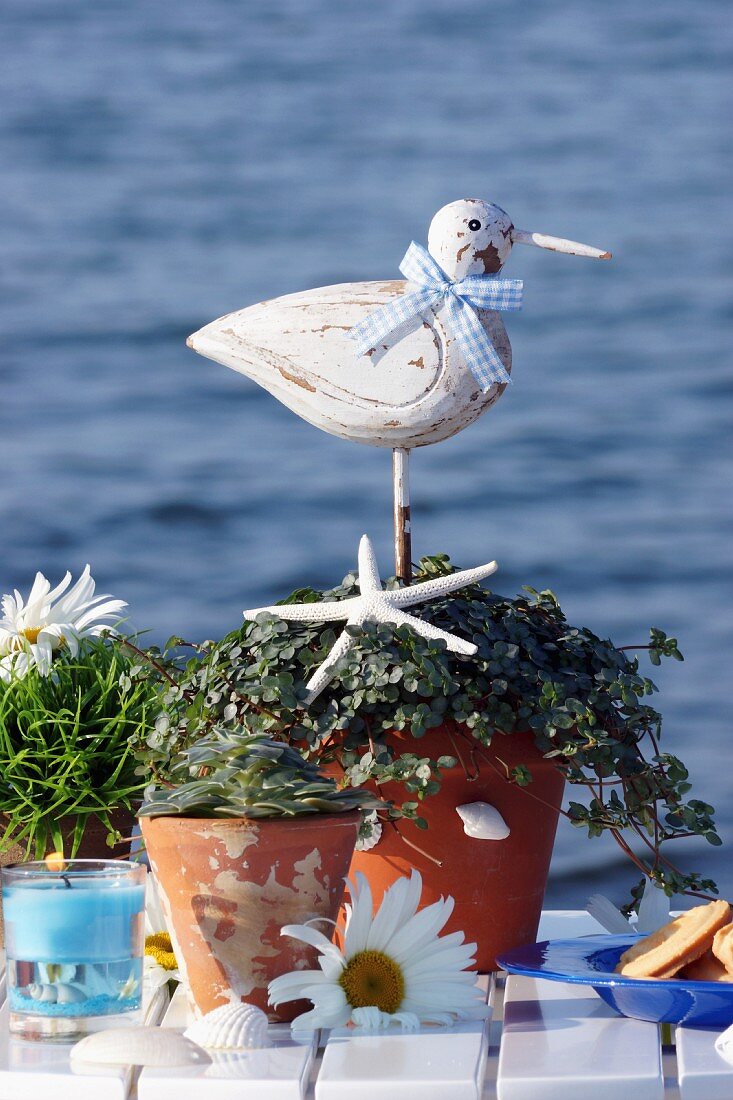 Verschiedene Pflanzentöpfe mit Deko Vogelfigur auf Tisch vor Seehintergrund