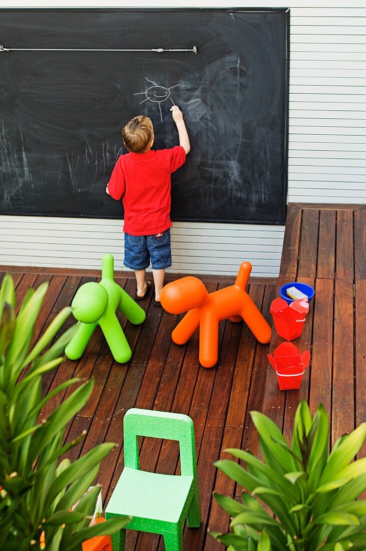 Terrasse mit Kinder Spielsachen auf Holzboden, kleiner Junge vor Schiefertafel beim Zeichnen