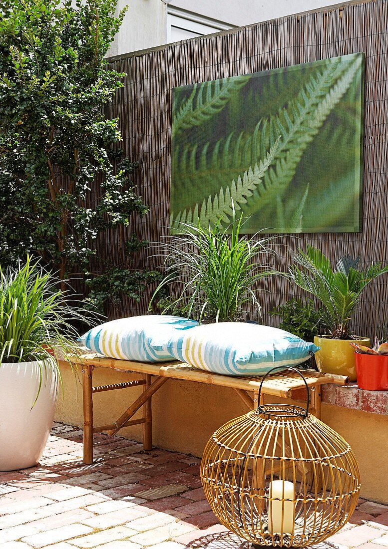 Wicker lantern on terracotta floor in front of bamboo bench with cushions and photo print on screen fence in courtyard