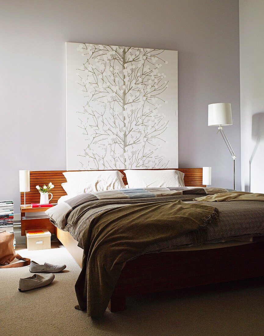 Gemälde mit stilisiertem Baum über dem hölzernen Kopfteil eines Doppelbetts vor grau getönter Wand; Designer Stehleuchte in der Ecke