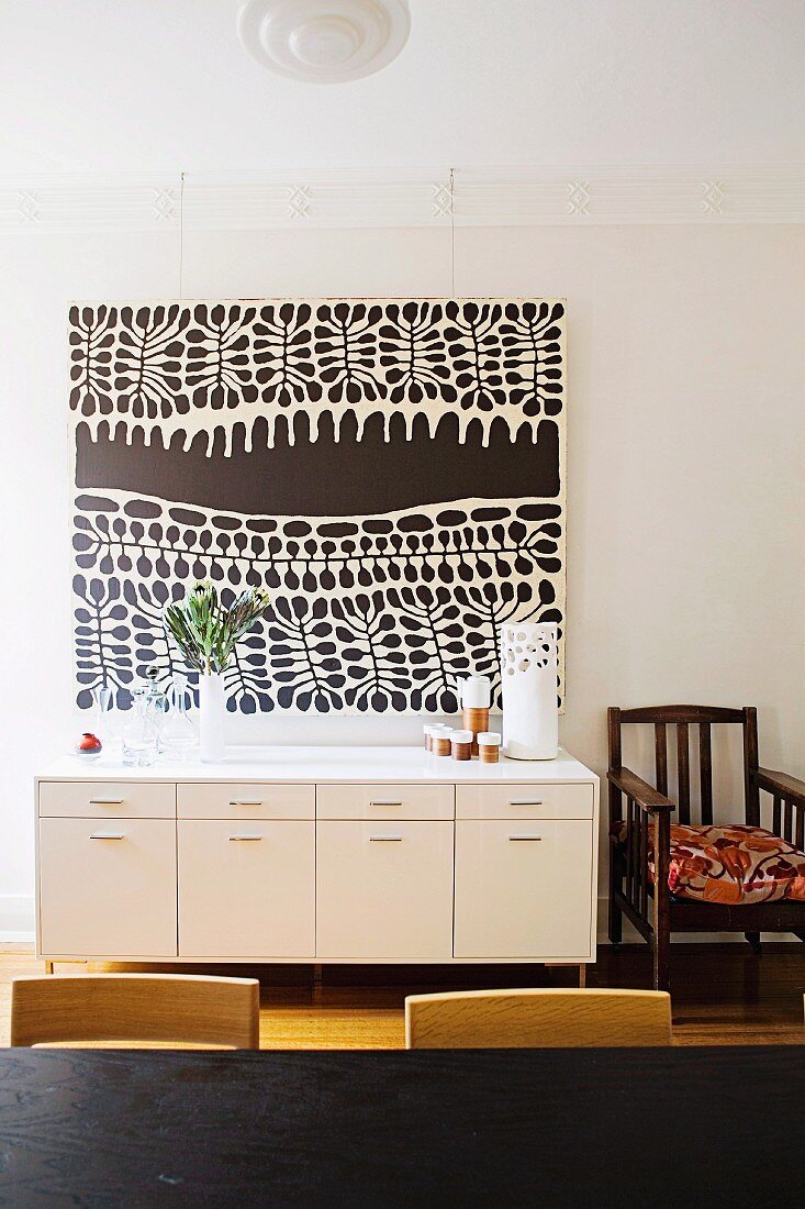 Modern black and white painting above white sideboard next to simple, wooden armchair with cushion