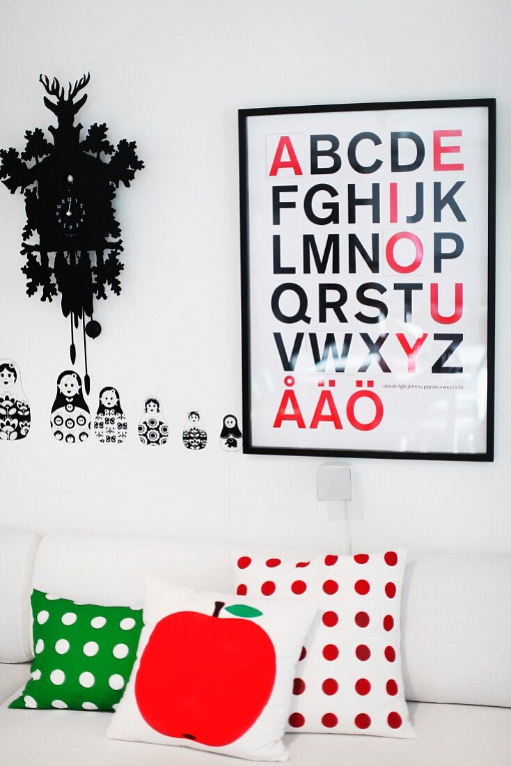 Kissen mit verschiedenen Mustern auf weisser Couch vor Wand mit gerahmtem Plakat neben Kuckucksuhr