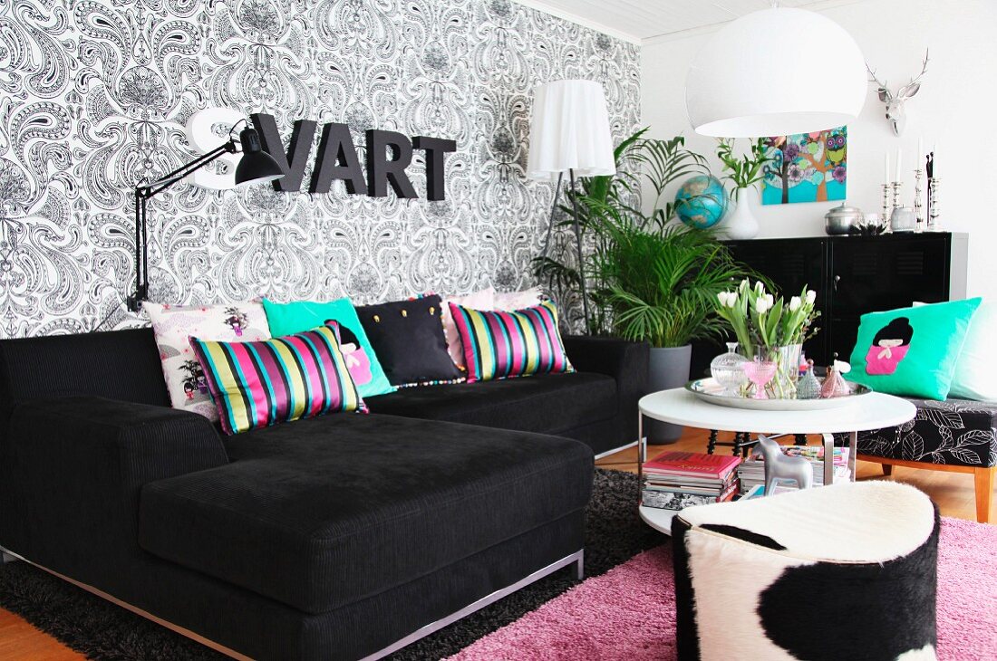 Black corner couch and pouffe around coffee table in living area with patterned wallpaper