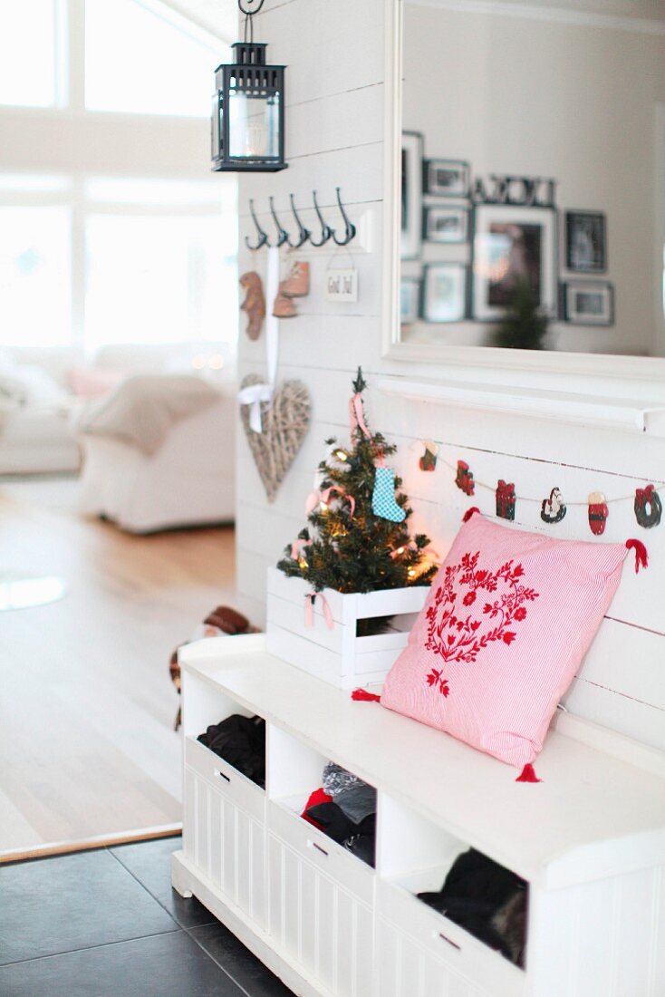 Pink cushion next to small, decorated Christmas tree in crate on white cloakroom bench in front of armchair in open-plan interior