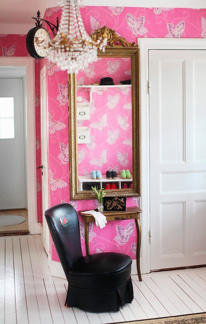 Black, leather lounge chair at console table and framed mirror on wall with butterfly-patterned wallpaper on pink background
