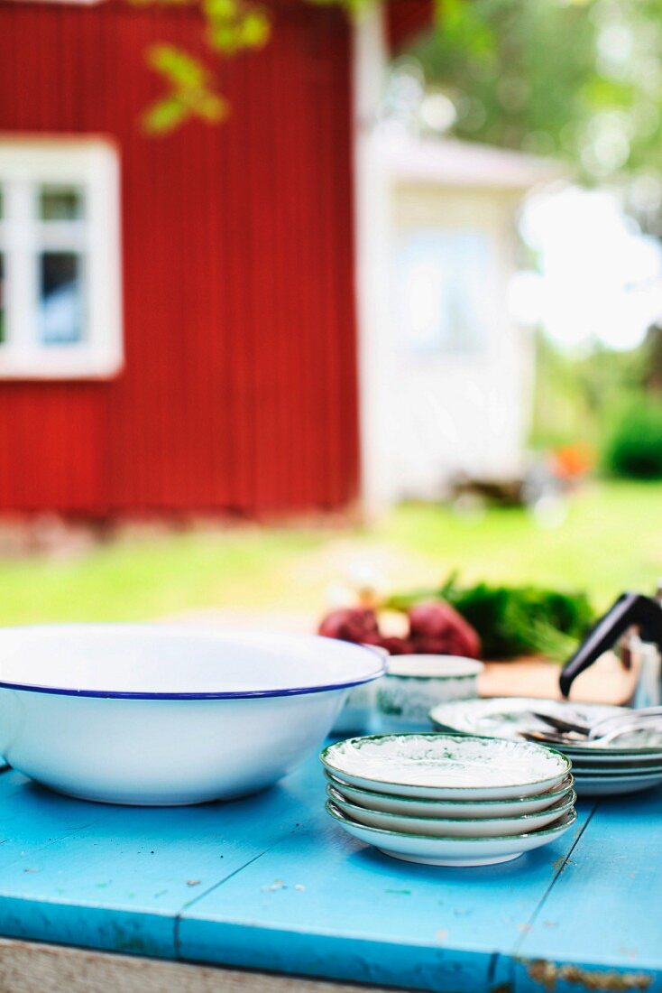 Tellerstapel und Emailleschüssel auf blau lackiertem Gartentisch, im Hintergrund rotes schwedisches Holzhaus