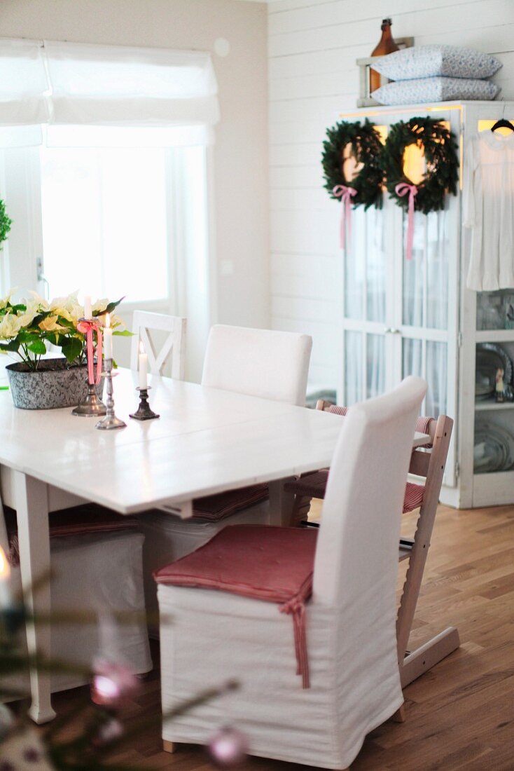 Table and chairs, some with white loose covers, in front of wreaths hanging on cupboard door