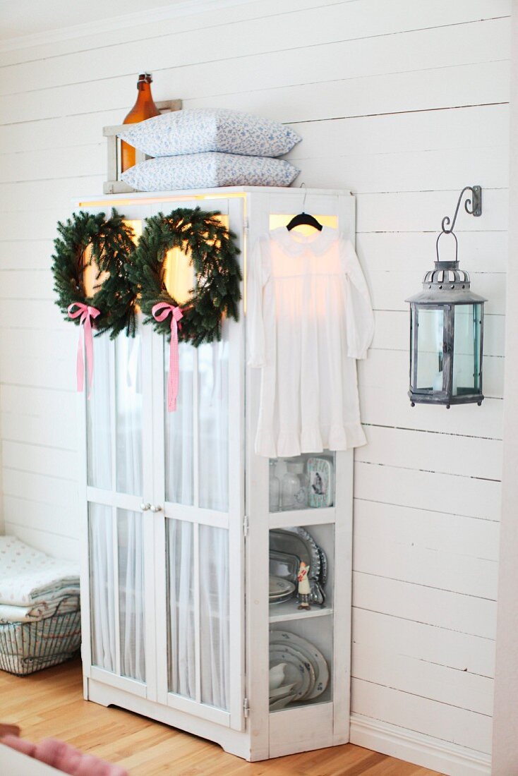 Christmas wreaths of fir branches and ribbons hanging on cupboard door in rustic interior with white wood cladding