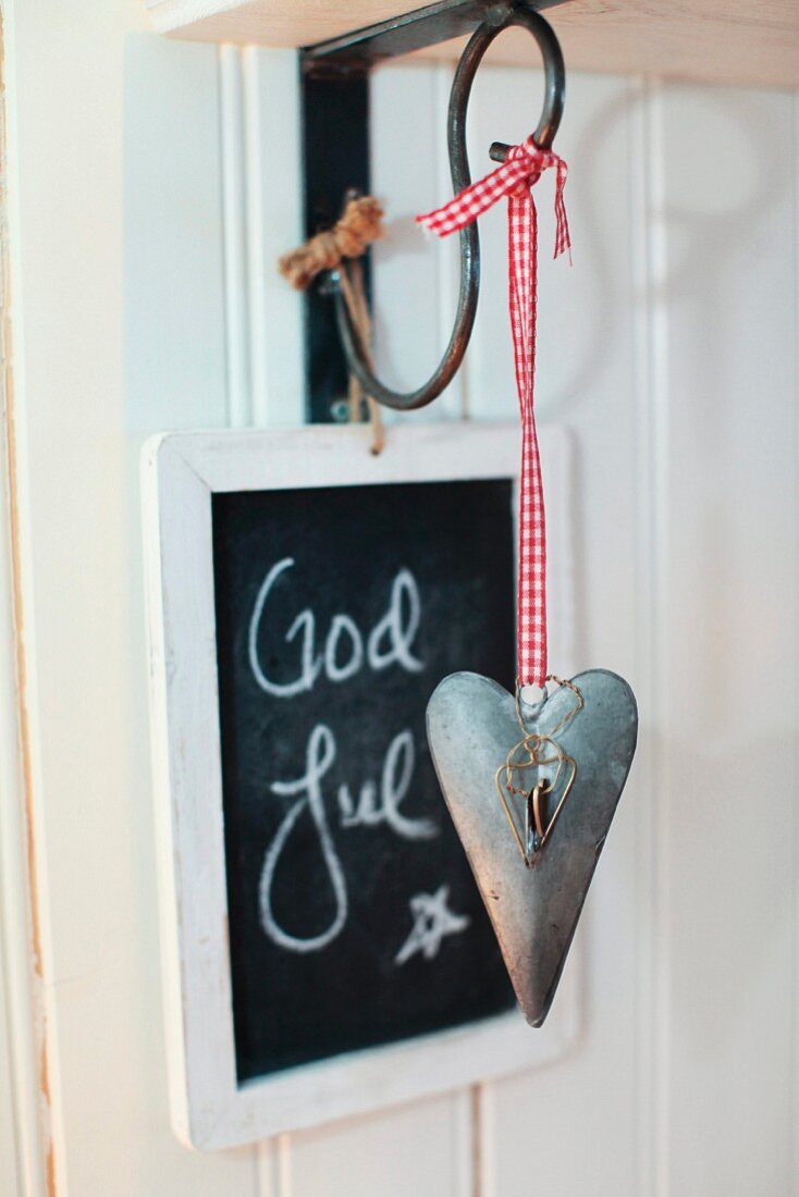 Heart-shaped pendant with red gingham ribbon hanging from wall bracket in front of Christmas greeting on blackboard
