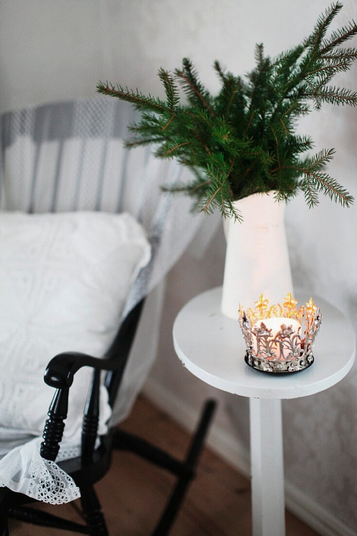 Ornate metal candle lantern in front of white water jug of fir branches on side table next to black rocking chair