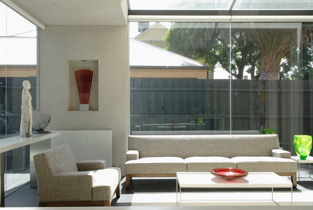 Seating area in glass extension with pale, designer sofa and armchair and colourful glass vessels on side tables; collectors' pieces in niche and on shelf to one side