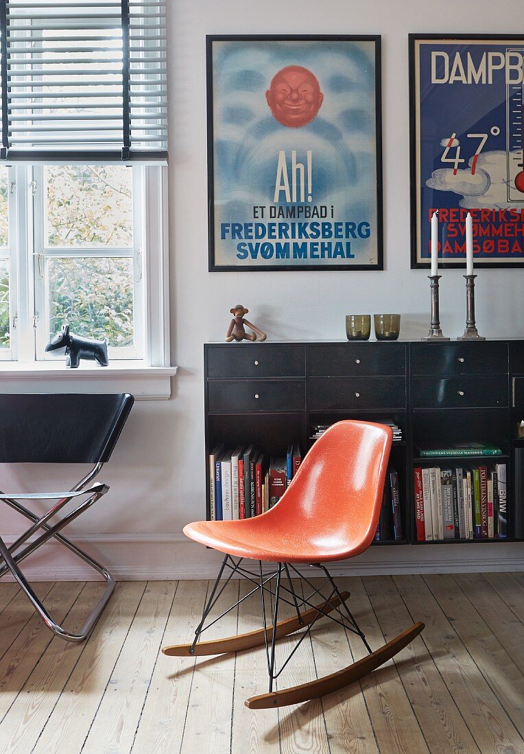 Charles Eames Schaukelstuhl mit orangeroter Sitzschale vor Sideboard an Wand montiert, darüber gerahmte Poster