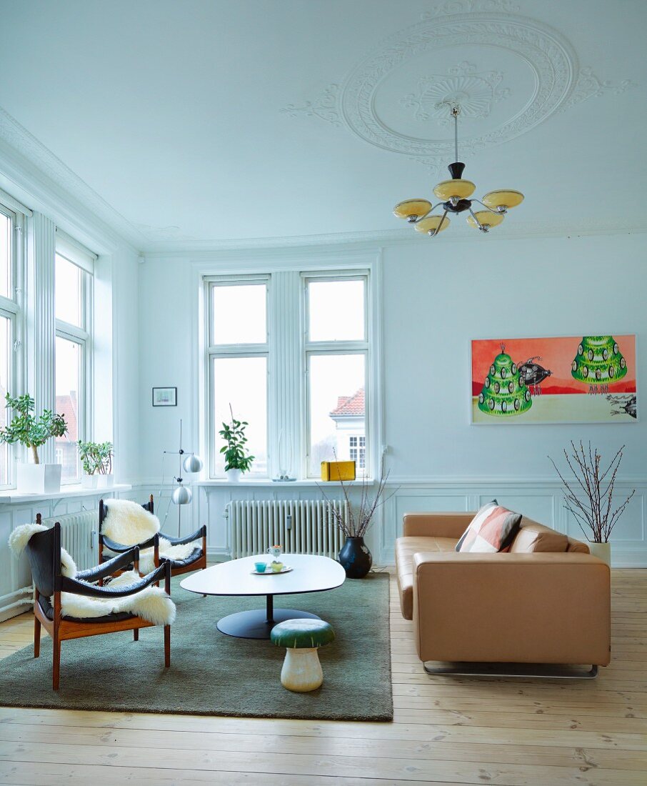 Sessel mit Holzgestell und weißem Tierfell, hellbraunes Ledersofa um Couchtisch mit amorpher Platte, in Wohnzimmer, Art Deco Hängeleuchte an Stuckdecke