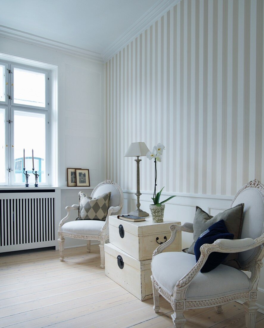 Stack of pale, vintage trunks flanked by Rococo-style armchairs against striped wallpaper in elegant interior