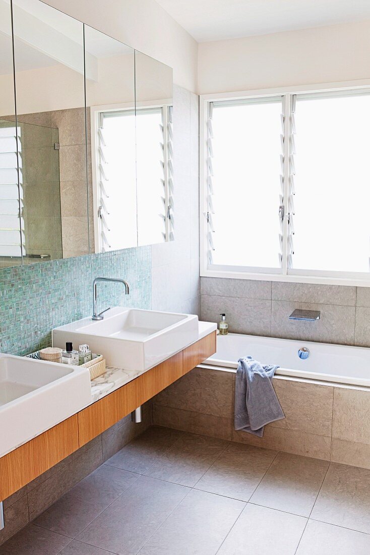 Modern bathroom with mirrored cabinet above twin sinks next to bathtub below window