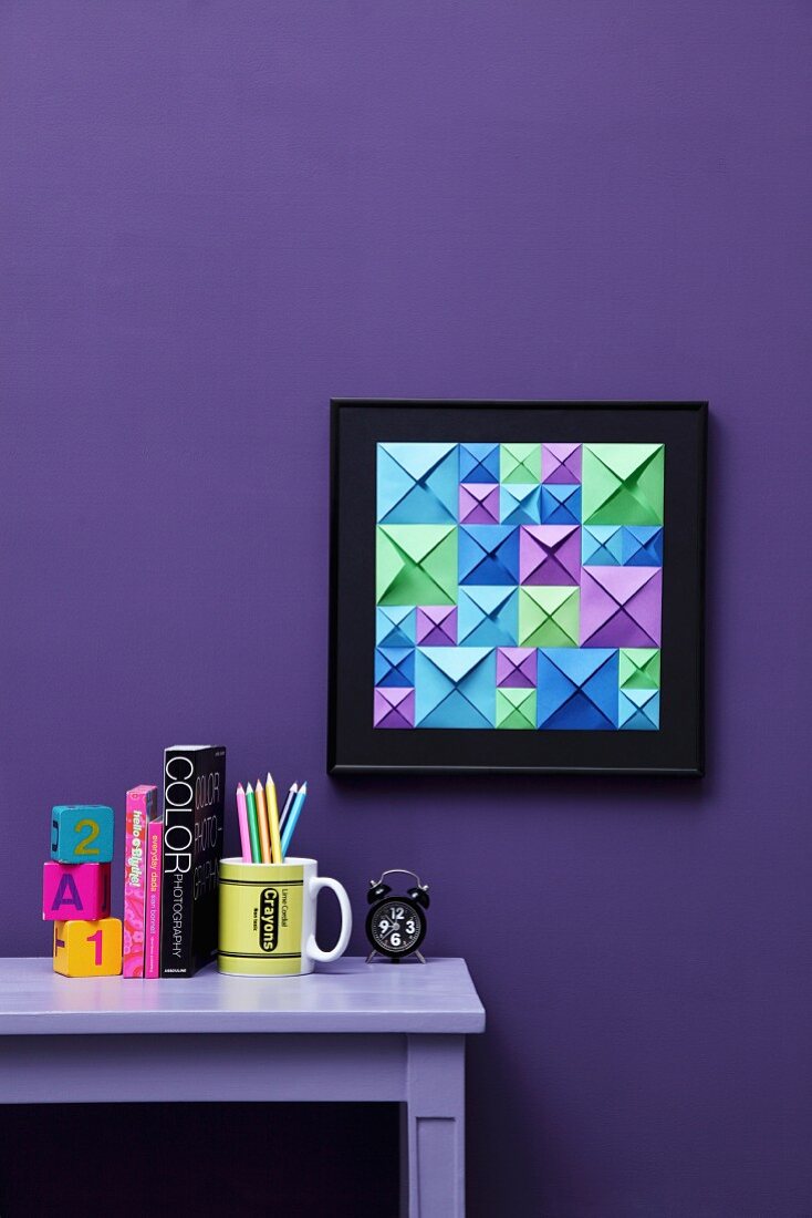 Hand-crafted, 3D-effect artwork made from blue, green and purple origami squares of different sizes on violet wall; matching ornaments on desk