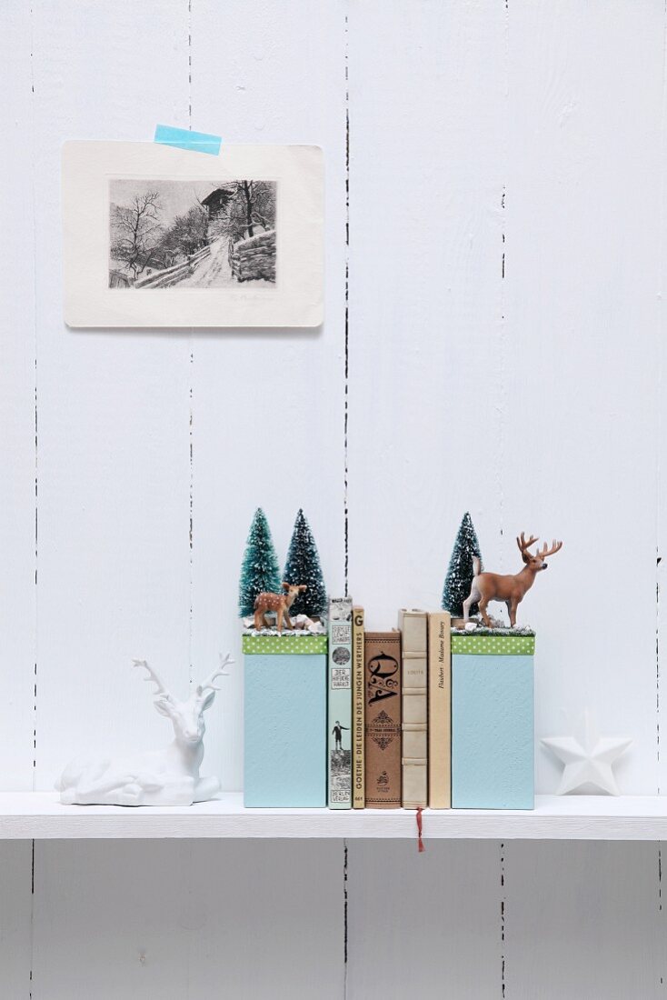 Selbstgebastelte Buchstützen mit Winter-Motiven, Miniatur-Rehe und Bäumchen-Deko auf Ablage vor weisser Holzwand