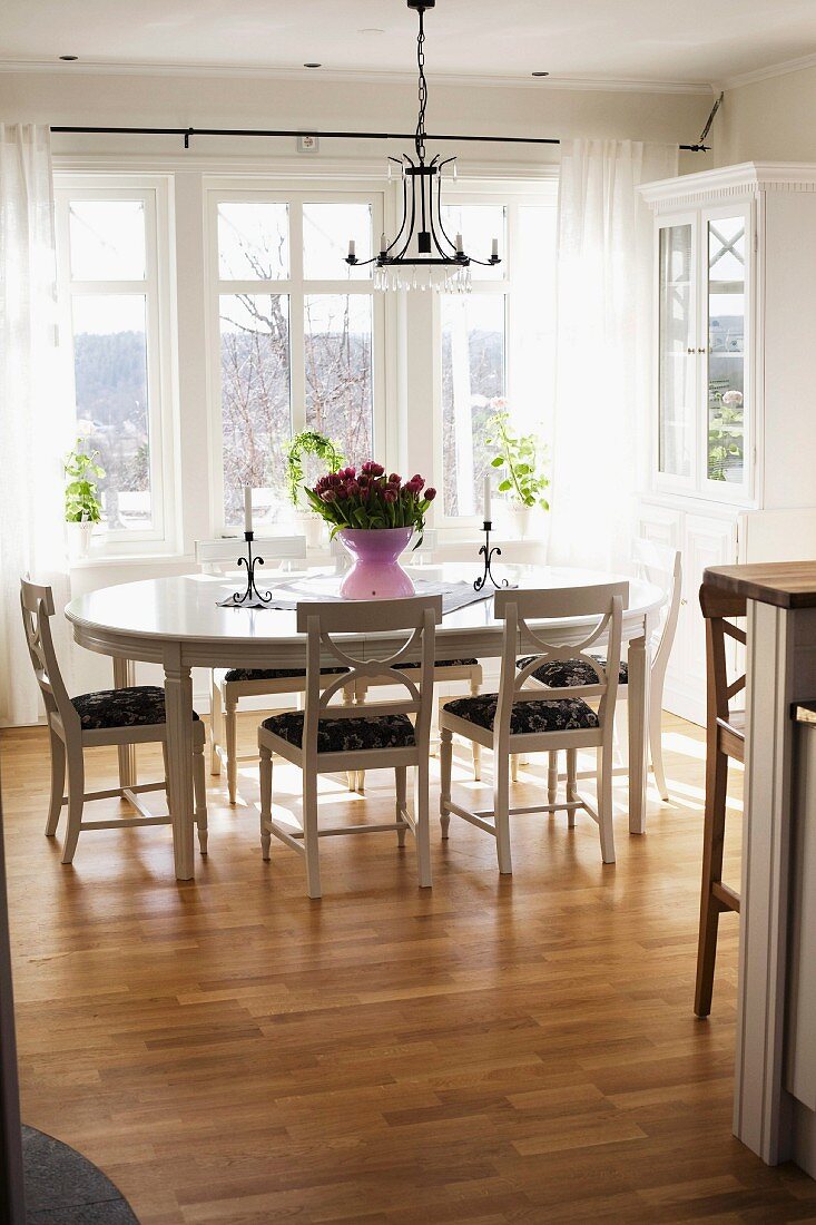 Ovaler Esstisch und Holzstühle in Weiß unter schmiedeeisernem Kerzenleuchter vor Fenster