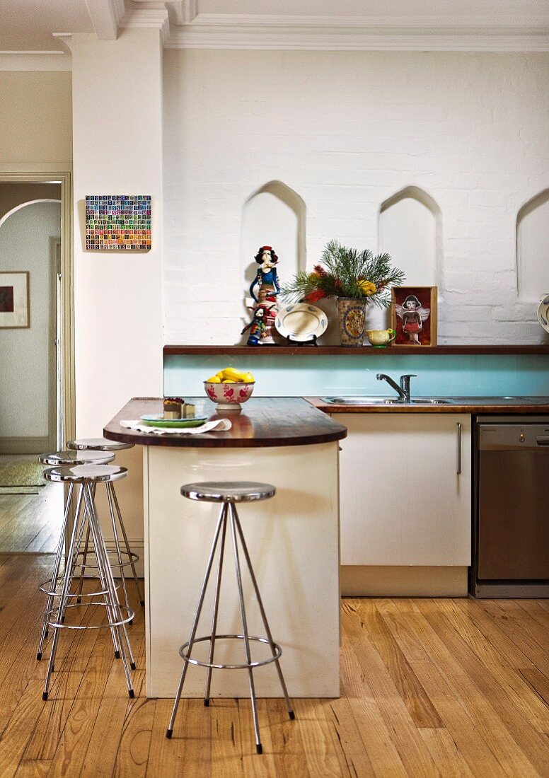 Retro-Barhocker aus Chrom vor Küchentheke auf Dielenboden, an Wand Holzablage unter kleinen Nischen mit Spitzbogen