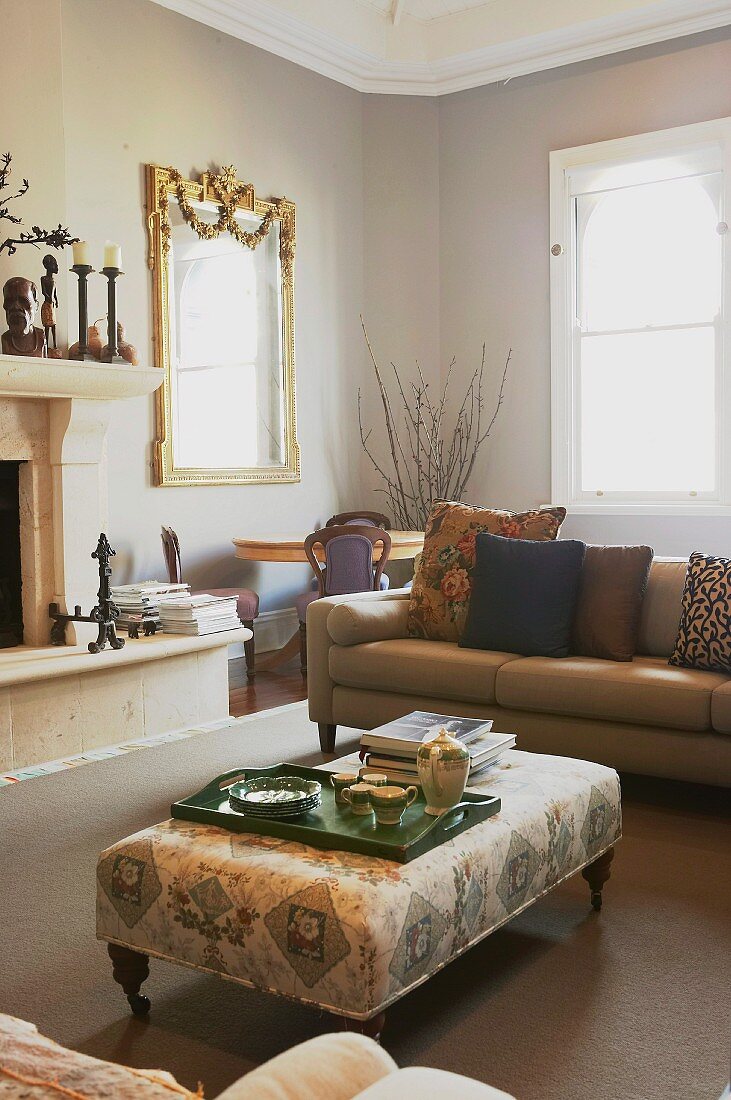Tea set on tray on ottoman and timeless sofa in front of fireplace; gilt-framed mirror on background
