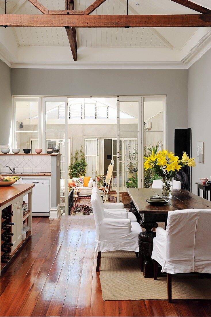 Antique table, loose-covered chairs and exposed roof structure in dining area of open-plan kitchen