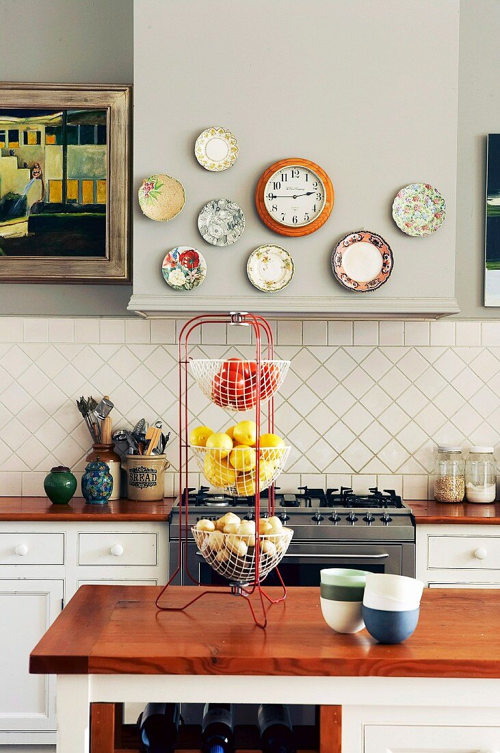 Decorative plates and wall clock on extractor hood above gas cooker in country-house kitchen with fruit and vegetables in 3-tier wire basket
