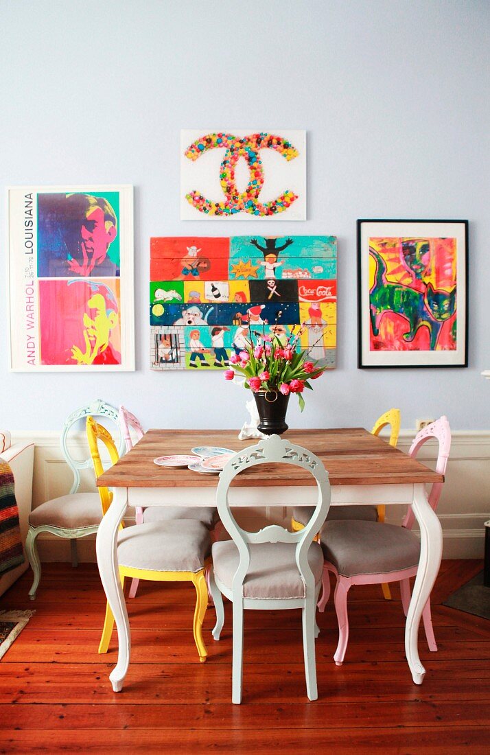 Essplatz mit farbig lackierten Antikstühlen und farbenfrohen Bildern an der lichtgrau getönten Wand einer Altbauwohnung