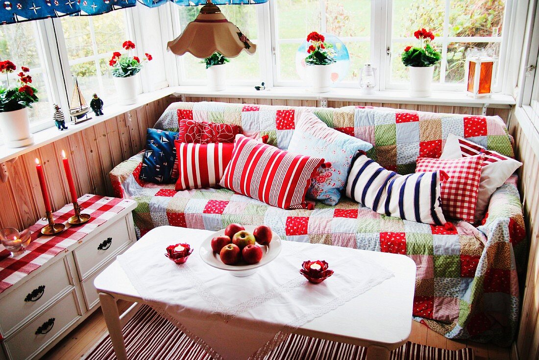 Gemütlicher Platz in der Loggia - weisser Tisch und Sitzbank mit Patchworkdecke vor Brüstungswand aus Holz, auf Fensterbank Blumentöpfe mit roten Geranien
