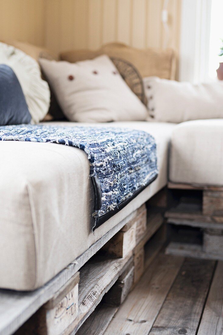 Close-up of DIY bench made from wooden pallets and pale seat cushions with rag rug and scatter cushions