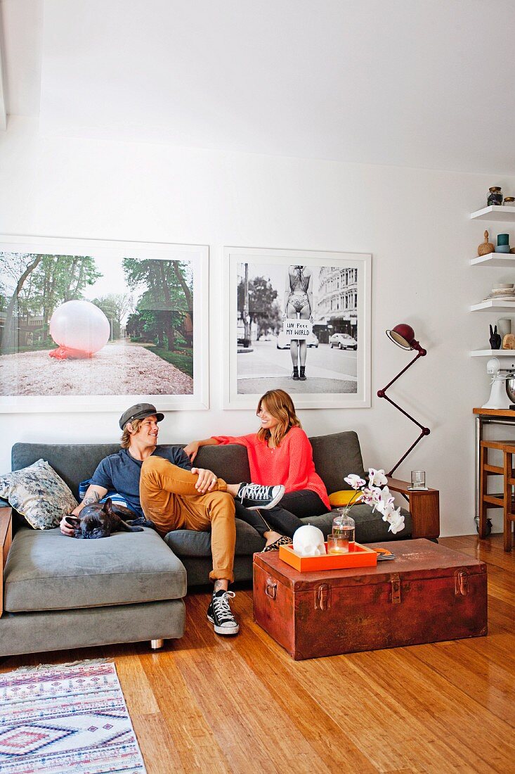 Junges Paar auf gemütlicher Polstercouch, davor schlichte Holztruhe auf Dielenboden, an Wand grossformatige Fotos