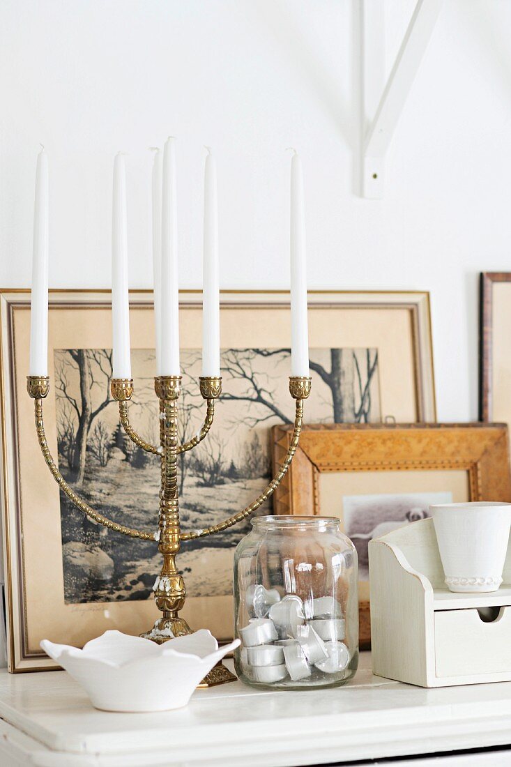 Multi-armed brass candelabra and jar of tealights in front of framed pictures on white shelf