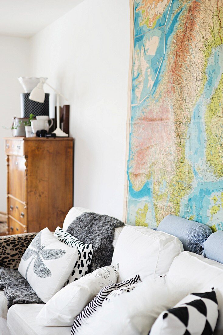 Various scatter cushions on white sofa in front of map of the world on wall and rustic wooden cabinet in background