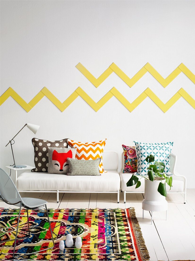 Colourful, patterned rug on white wooden floor in front of upholstered bench with scatter cushions of various patterns; yellow zigzag pattern on wall