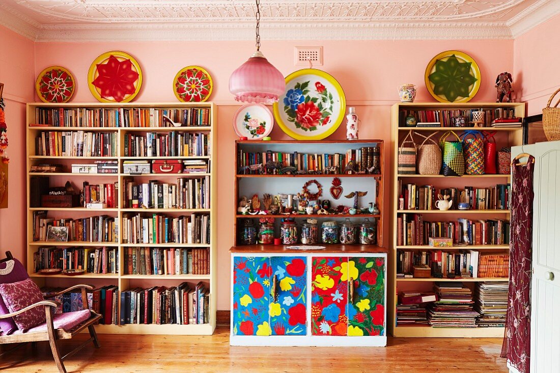 Library area with pink-painted walls and colourful floral dresser between full bookcases in collector's apartment