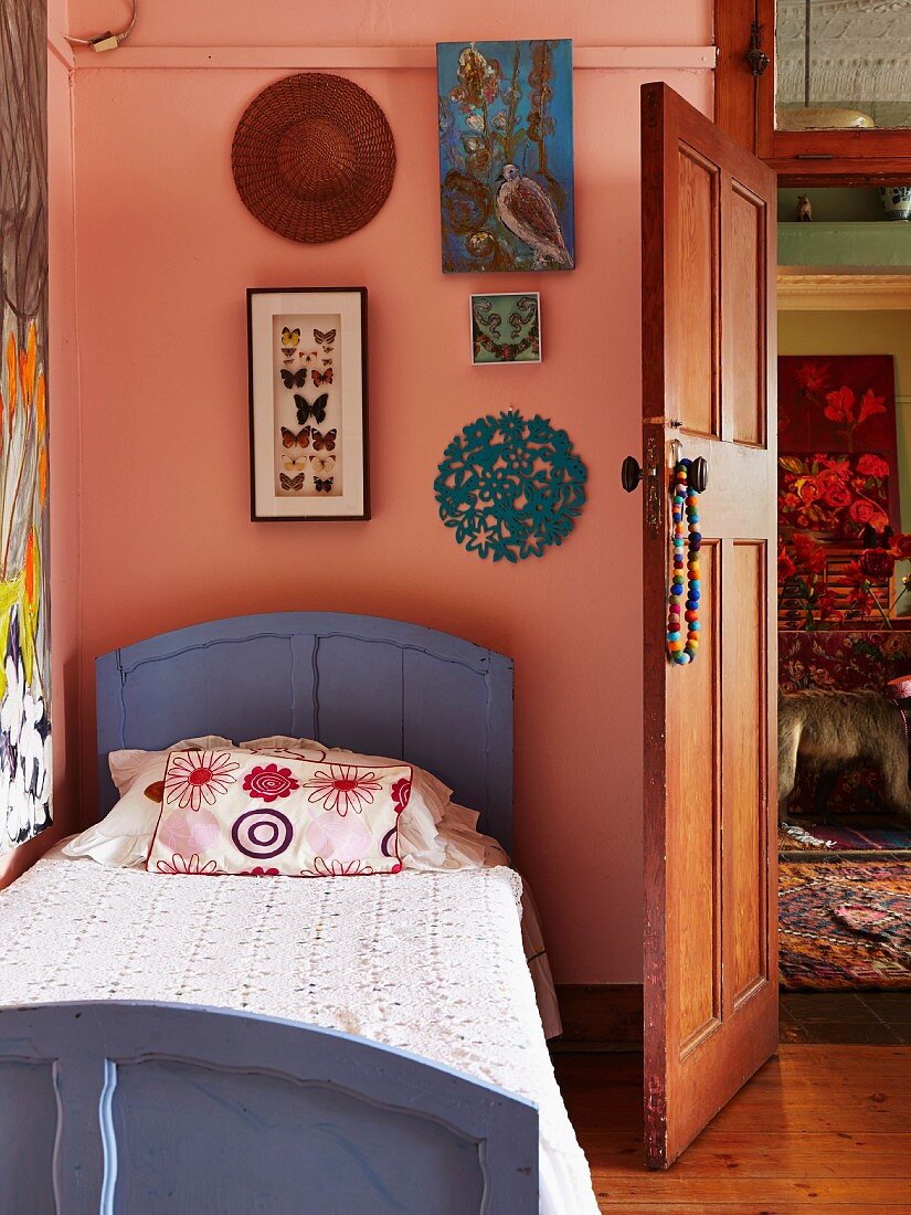 Violett lackiertes Holzbett vor rosafarbener Wand mit gerahmter Schmetterlingsammlung und Bildern, seitlich offene Holztür