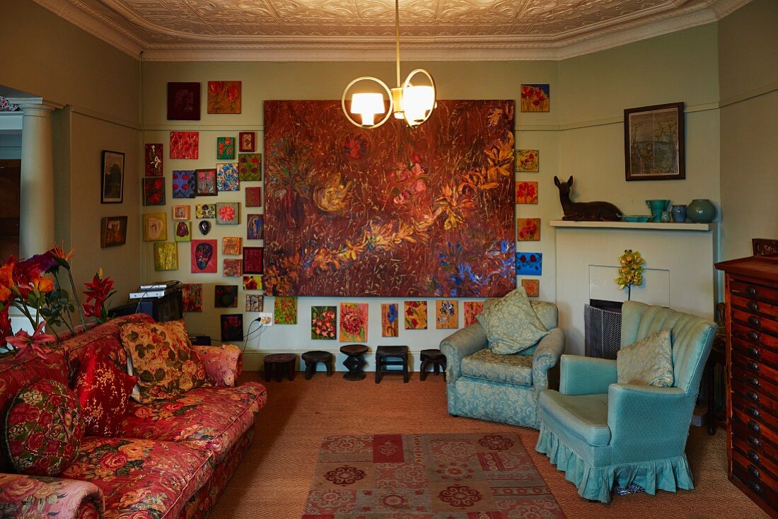 Polstersofa mit Vintage Blumenmuster und Sessel mit hellblauem Bezug in künstlerischem Wohnraum, an Wand grossformatiges Gemälde inmitten vieler kleiner Bilder