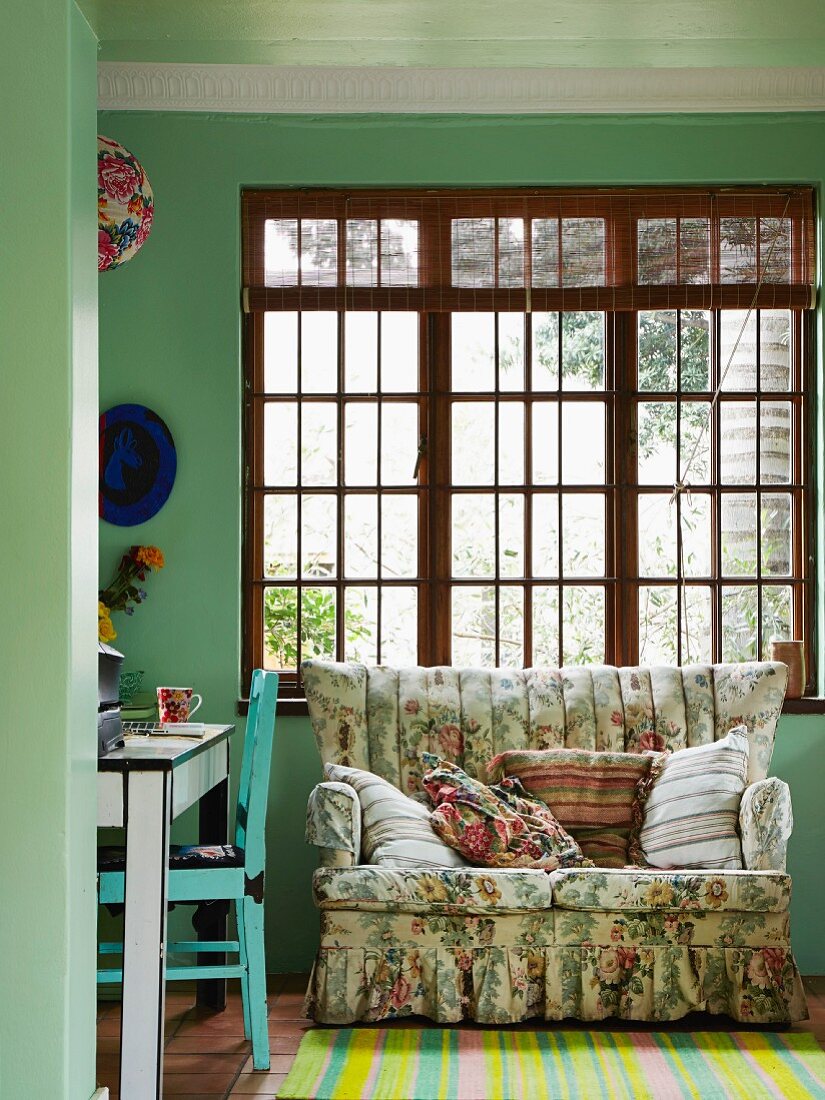 Gemütliches Sofa mit Vintage Blumenmuster vor Sprossenfenster aus Holz, davor gestreifter Teppichläufer mit kräftigen Farben in grün getöntem Wohnbereich