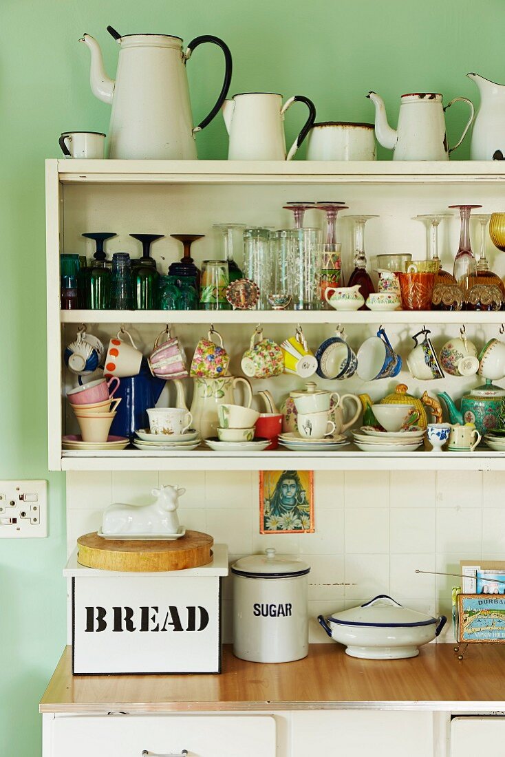 Crockery and collection of vintage enamel jugs on retro kitchen shelves on pale green wall