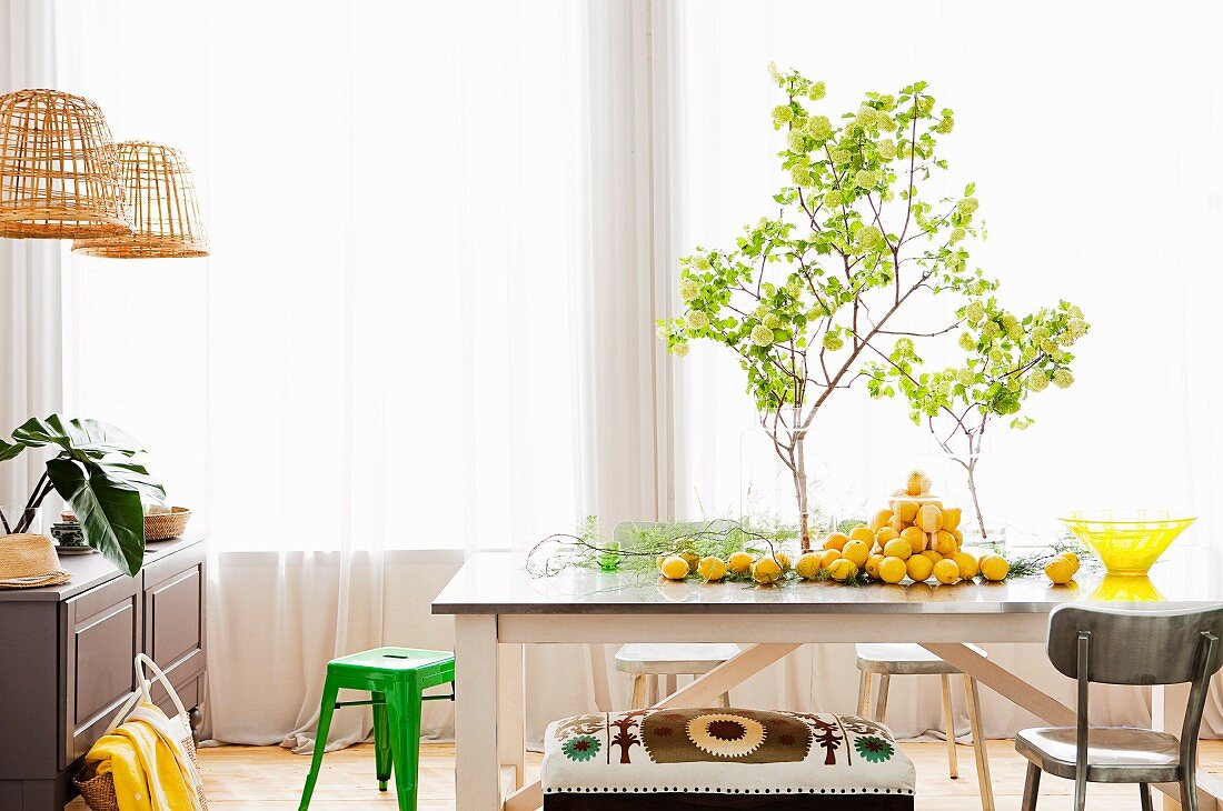 Blätterzweige in Vase und Zitronen auf ländlichem Holztisch verteilt, dahinter Fenster mit luftigen Vorhängen