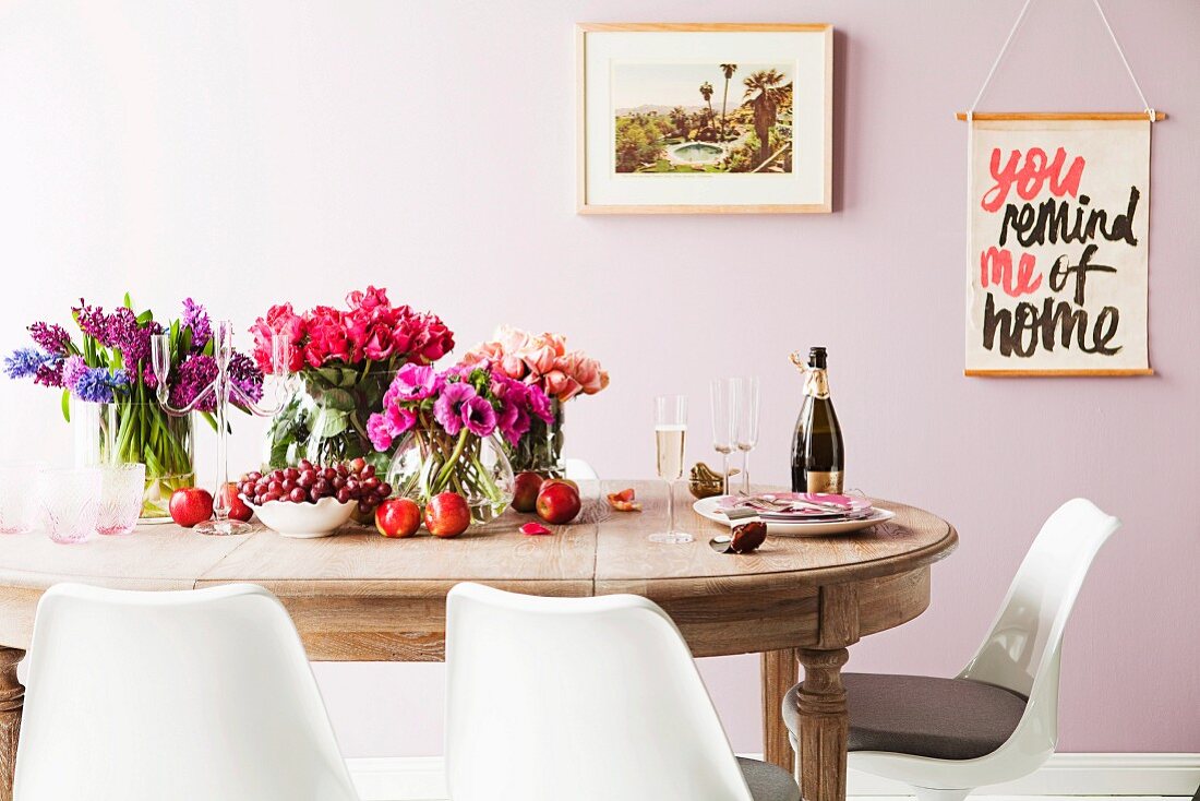 weiße Schalenstühle im Retro Stil um antikem Holztisch mit Blumensträussen in Glasvasen und roten Früchten, vor pastellrosafarbener Wand