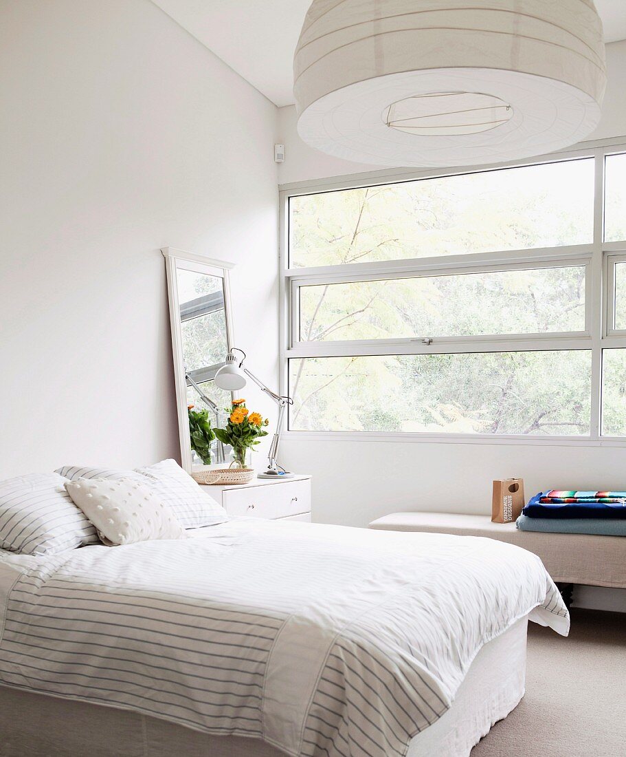 Bright, white bedroom with wide ribbon window and tall mirror leaning against wall on top of bedside cabinet