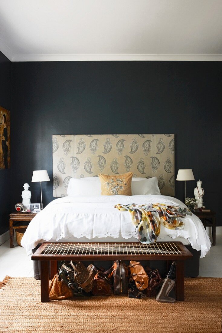 Doppelbett mit hohem, gepolstertem Kopfteil vor schwarz getönter Wand, im Vordergrund an Bettende Sitzbank mit Geflecht darunter aufgereihte Handtaschen