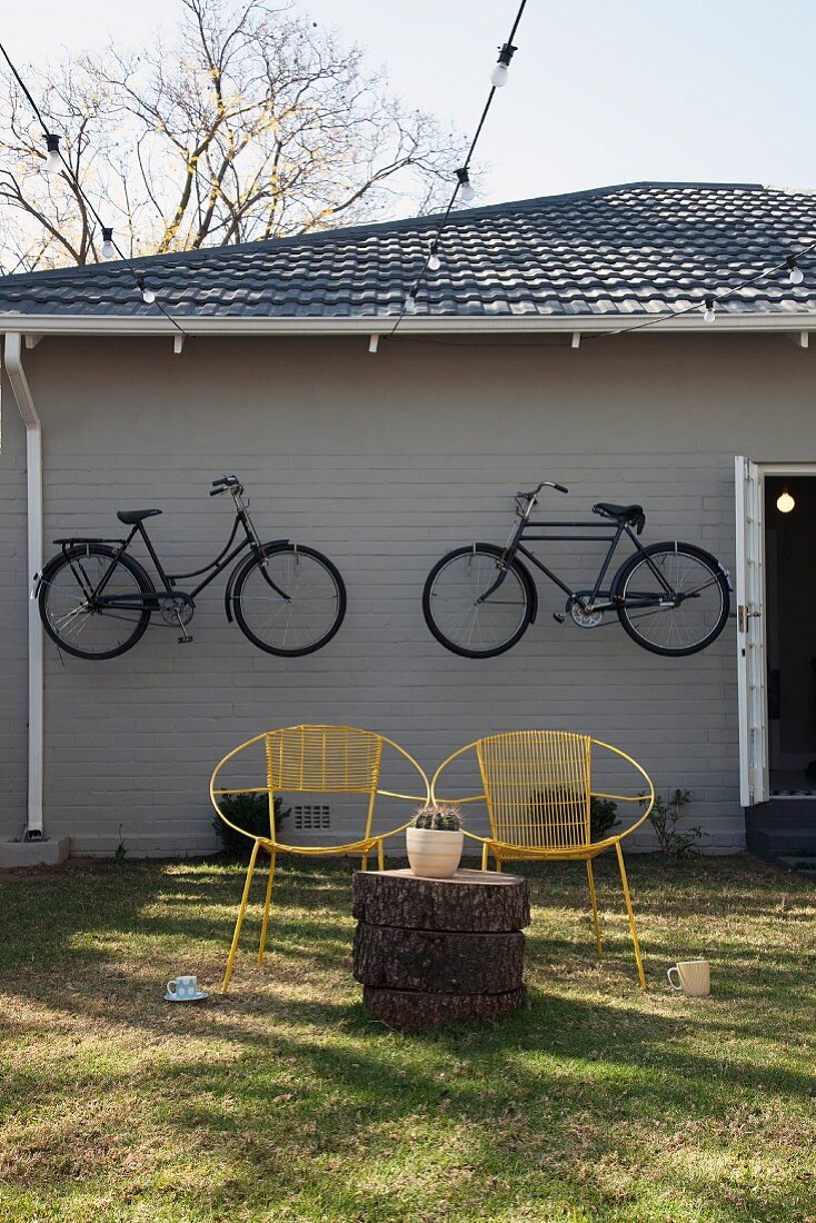 Gelbe Vintage Drahtstühle mit Baumstammtisch vor grauem Holzhaus mit aufgehängten Fahrrädern an der Fassade